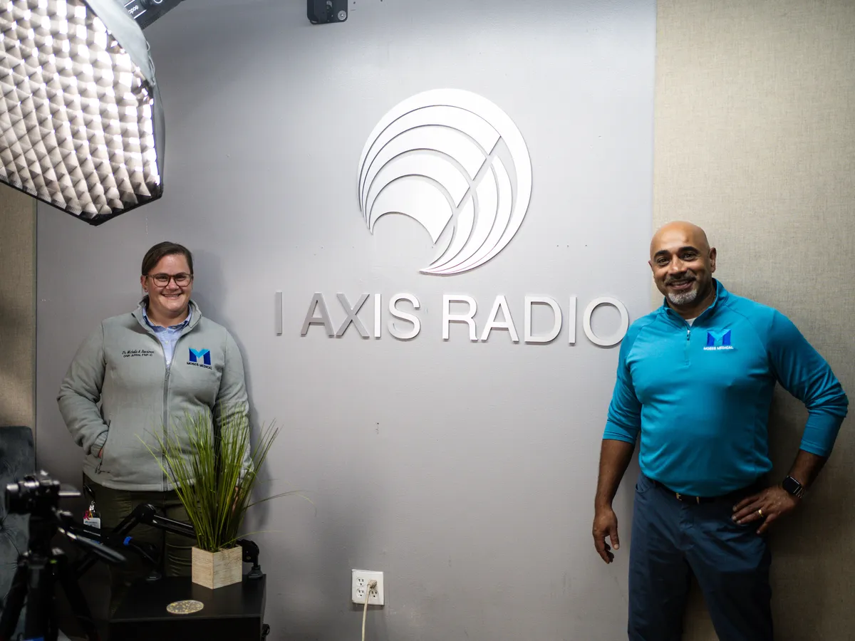 Dr. Michelle Benrimon in a gray Moses Medical jacket and John Johnson in a blue Moses Medical shirt standing by the I Axis Radio sign