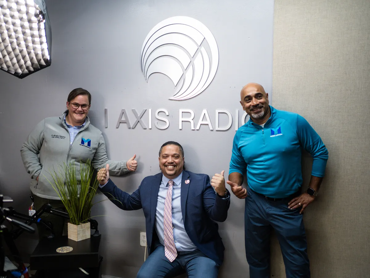 Dr. Benrimon and John Johnson by the I Axis Radio sign with the show's host seated in front, giving a thumbs up