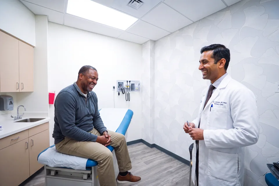 A man seated on an exam table laughs with a clinician in a white coat during a visit